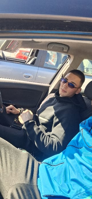 A young adult male is seated in the front passenger seat of a dark-interior vehicle. He has short hair and wears blue-tinted oval sunglasses, a dark hooded tracksuit jacket with a lighter stripe on the left sleeve, and dark trousers. A dark-colored smartphone is positioned on his lap near his right hand, which is loosely fisted. To his right, on the seat, a bright blue garment with black zipper details is visible. Strong sunlight enters the vehicle from the right, creating shadows. Through the passenger window, reflections and portions of other parked vehicles are discernible, specifically a red car and a light grey/silver car. The specific location within the city of Katina, Bulgaria, is not discernible from the visual information provided.