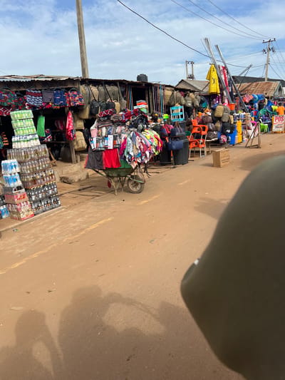 The image depicts a busy street market in Markurdi, Nigeria. Stalls line the street displaying various goods, including clothing, bags, and beverages. A cart laden with merchandise is visible, and there are stacks of packaged drinks on the ground. The environment suggests an active commercial area.