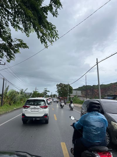 The image captures a traffic scene on a road in Lampisang, Indonesia, under a cloudy sky. A white SUV with license plate "BK 1209 XAE" is prominent in the foreground, facing away from the camera. Several motorcycles and other vehicles are visible in traffic ahead of the SUV. On the right side of the road, a person wearing a blue patterned shirt and a black helmet is riding a motorcycle. The surrounding environment includes vegetation, utility poles with numerous wires, and a rocky hillside in the background. The road appears to be wet, suggesting recent rain.