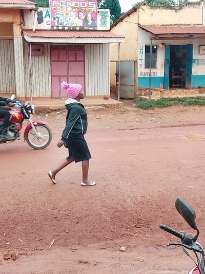 A woman in a pink knit hat and black skirt walks across a dirt road. She is wearing a black jacket. The road is in front of a building that says "PENDO'S COLLECTION" in large letters. The building has a pink facade with white trim. There is another building behind the woman that has a sign that says "UNITY KINYOZI" in large letters. It has a blue and white facade with a white trim. To the left of the woman is a motorcycle parked on the side of the road. There is a small amount of vegetation in the foreground. The location is Kivaywa, Kenya, at the coordinates 0.562, 34.805.