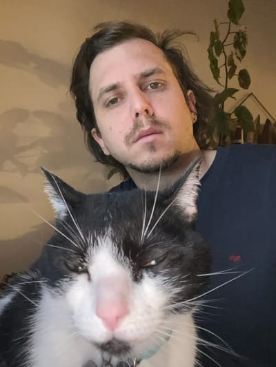 An indoor close-up image features a person and a cat, oriented sideways. The human subject, a man with shoulder-length dark hair, light-colored eyes, and stubble on his chin and upper lip, wears a dark blue shirt. He gazes directly at the camera with a neutral expression.
To his right, and closer to the camera, is a black and white tuxedo cat with prominent white whiskers and a pink nose. The cat's eyes are partially closed, suggesting a relaxed or sleepy state. It appears to be wearing a light blue collar with a metallic tag.
In the blurred background, a large-leafed green plant is visible behind the man's head. The wall behind them is a plain, light tan or beige color. A glimpse of a framed object can be seen in the upper right. A small, red, indistinct object is present in the dark blue area of the background behind the cat's head.
The scene depicts a candid moment between the man and the cat, possibly a selfie. The lighting is soft and even, typical of indoor conditions. The location context provided is Ushuaia, Argentina. No visible text is present in the image.