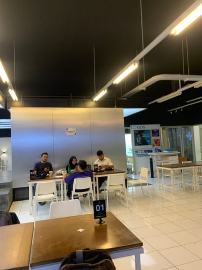 The image shows a seating area in a cafe located at Jalan Letjen S. Parman, Kota Malang, Jawa Timur, Indonesia (latitude: -7.941, longitude: 112.608). Four people are seated at a table in the foreground, two men and two women. The men are wearing casual clothing, and one of the women is wearing a headscarf. They are all looking down at their phones or devices. There are white chairs and a brown table. The table is numbered "03". There is another brown table to the left, with number "01" on it. A backpack sits on this table. The cafe has white tiled floors, and there are white chairs and tables scattered throughout the seating area. The cafe's ceiling is black and has several rows of recessed lights. An IQOS advertisement sign is visible on the right side of the image, behind a counter. There is a window to the right of the counter, and a person's legs are visible through the window. The walls are made of metal paneling, and they are painted in a light gray color. The room is well-lit. The cafe is relatively empty, with only the four people seated at the table in the foreground. There are no other people or animals present in the image.