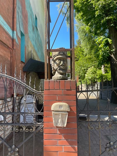 The image shows a sculpture of a bearded head mounted on a brick pillar, situated in front of a metal gate. The sculpture is made of stone and has a weathered appearance. It is located in the city of Kaliningrad, Russia, specifically at the entrance to a shop that sells antique items and souvenirs. The building behind the gate is decorated with a colorful mural featuring geometric patterns and windows. The environment is sunny with lush green trees visible in the background, indicating a daytime scene. The gate itself is ornate, made of black wrought iron with intricate scrollwork and floral motifs. A sign is visible behind the gate, advertising the shop and its offerings.