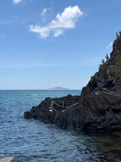 The image depicts a coastal scene in Phú Lương, Vietnam. In the foreground, dark, layered volcanic rocks form a rocky shoreline. The sea is a vibrant blue, with gentle waves lapping against the rocks. A rope barrier, supported by white posts, guides visitors along the edge of the rocks. In the distance, the vast expanse of the sea stretches to the horizon, meeting a clear blue sky dotted with a few fluffy white clouds. A small, hazy island is visible in the far distance. The overall impression is one of natural beauty and tranquility, with hints of human presence through the rope barrier and a small buoy in the water.