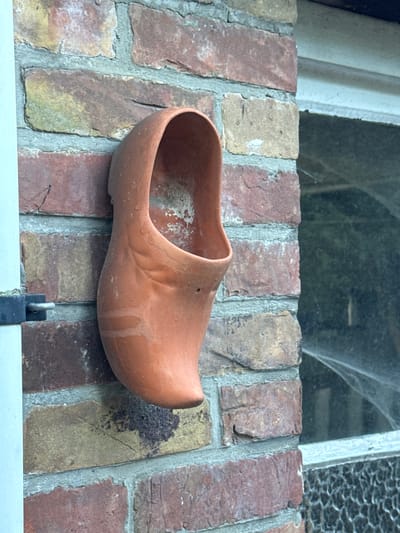 The image shows a terracotta clog, likely a planter, mounted on a brick wall. The clog is oriented vertically, with its opening facing upwards and to the left. It appears to be empty and shows signs of weathering, with some white residue and streaks on its surface. The brick wall is constructed with irregular, reddish-brown and yellow bricks, mortared with light grey grout. To the right of the clog, a window pane is visible, partially obscured by shadows and what appears to be cobwebs. The window frame is light colored, possibly white or pale blue. A black metal latch is attached to the left side of the wall. The overall scene suggests an outdoor setting, possibly a garden or the exterior of a building. There are no people or animals visible in the image. The image does not provide specific geographical markers to pinpoint a location within Bruges, Belgium.