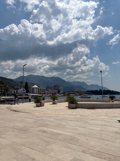 The image captures a scenic waterfront promenade in Tivat, Montenegro, under a partly cloudy sky. In the foreground, a wide paved area, likely a public square or walkway, is laid with large, light-colored rectangular stone tiles. The stones are arranged in a staggered pattern.
To the left of the foreground, the promenade curves towards the water. Several people are visible on or near the water's edge. One person in a dark top and light shorts is walking along the promenade, and other individuals, some appearing to be children, are near the water.
The middle ground features a body of water, likely the Tivat Bay, with several small boats and yachts anchored or moving. Along the waterfront are buildings with tiled roofs and balconies, typical of coastal Mediterranean architecture. There are also planters with palm-like plants lining a raised, curved wall. Two tall lampposts are present, one closer to the left and another further to the right, illuminating the area.
The background is dominated by a range of lush green mountains that frame the bay. The sky above is a mix of bright blue patches and large, dramatic white and grey cumulus clouds, suggesting dynamic weather conditions. The overall impression is a vibrant, sunlit coastal scene.