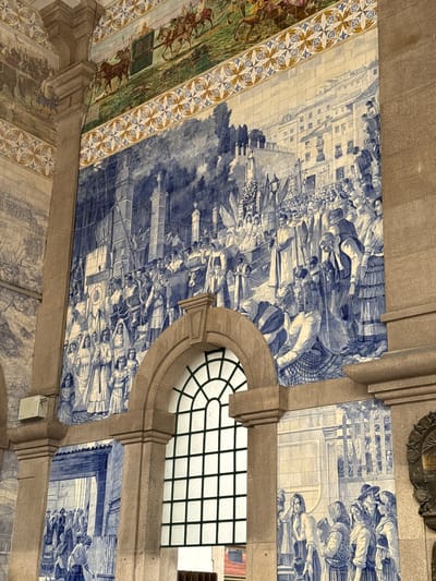 This image captures a section of the azulejo tile panels adorning the São Bento train station in Porto, Portugal. The primary focus is a large, detailed mural rendered in blue and white tiles, depicting a historical scene of a procession or celebration. Numerous figures, dressed in traditional clothing of various eras, are shown moving through a cityscape with architectural elements such as buildings, fences, and lampposts. Some individuals are holding flags or banners, suggesting a significant event. The composition is dense with people, with a large crowd filling the foreground and extending into the mid-ground. Above this central panel, another tiled section features a smaller, more colorful depiction of horse-drawn carriages and riders in a pastoral setting, bordered by an ornate geometric pattern. To the left and below the main mural, other azulejo panels continue the narrative with different scenes, including what appears to be a battle or military activity, and more street scenes with groups of people. The tiles are framed by substantial stone pillars and archways, including a large arched window with a grid pattern. The lighting suggests an indoor environment, likely the grand hall of the train station, with light illuminating the tiled artwork.