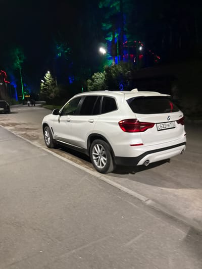A white BMW X3 SUV is parked on the right side of a paved road at night. The car is positioned at an angle, facing away from the camera and towards the right. The license plate is visible, reading "C623YE 174". In the background, trees are illuminated with colored lights, predominantly green and blue, with some red lights visible further in the distance, suggesting a decorative or festive lighting display. A street lamp is visible on the left side of the image, casting light onto the road. The overall environment appears to be an outdoor area, possibly a park or a recreational space, in the city of Turgoyak, Russia. There are no people clearly depicted in the immediate vicinity of the car, though faint figures can be seen further down the road in the background. The road surface is asphalt, and there is a curb separating the road from a lighter-colored surface, possibly a sidewalk or a path, to the left.