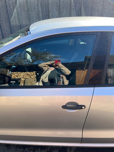 The image displays the front and rear passenger doors and window of a silver metallic car, parked outdoors in Wüstenrot, Germany, under bright sunlight. A reflection is prominently visible in the front passenger window. An adult, wearing a dark top and a light-colored cardigan, holds a red smartphone with both hands, appearing to photograph the reflection. An infant, dressed in dark clothing and partially covered by a light-colored wrap, is cradled or carried by the adult. Behind the reflected figures, the reflection shows a neatly stacked woodpile, a black box marked "OWA LAND ROVER," a building with a dark facade and a red-tiled roof, and bare tree branches against a blue sky. The car's front passenger door features a black handle with a visible keyhole. The actual background behind and above the car is a tall, weathered corrugated metal fence or wall. The ground beneath the car is dark asphalt.