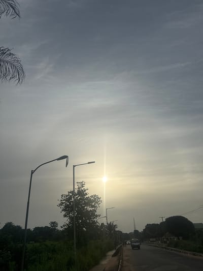 The image captures a street scene in Awka, Nigeria, likely during the late afternoon or early morning, given the diffused sunlight. The sky is overcast with a soft, pale glow emanating from the sun, which appears as a bright, star-like orb low on the horizon, casting a gentle haze over the scene.

Two streetlights are prominently featured in the foreground. The one on the left is taller and has a curved arm, while the one on the right is shorter and more angular. Both are silhouetted against the bright sky. Beyond them, a road stretches into the distance, curving slightly to the right. A few vehicles are visible on the road, including a white minivan in the center and a dark-colored car further down. The road has a paved surface with a white line marking the edge.

To the left of the road, there is a narrow, paved walkway bordered by tall, green grass and vegetation. Several trees are scattered throughout the landscape, with lush green foliage. In the background, more trees and some buildings can be seen, partially obscured by the haze. A tall transmission tower is visible in the far distance, to the right of the road.

On the right side of the road, there is a low concrete wall or curb, with more vegetation and trees behind it. The overall impression is of a semi-urban environment with ample greenery. The lighting suggests a time of day with soft shadows and a muted color palette.