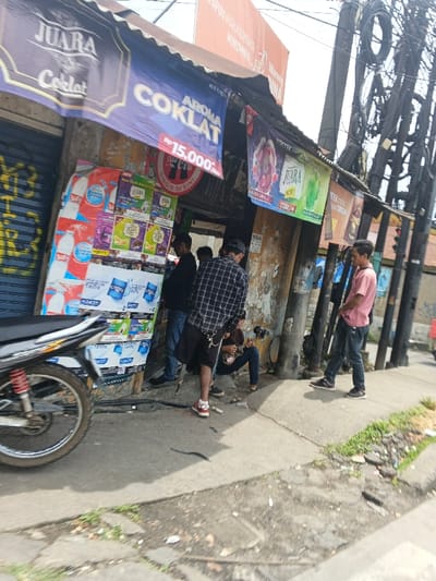 A street scene captures a small shop with a motorcycle parked outside. Banners advertise "JUARA Coklat" and "AROMA COKLAT," with a price of Rp15,000. People are standing and sitting nearby. The shot is taken during the day, with overhead wires and utility poles visible in the background. Debris is scattered on the curb near some greenery.