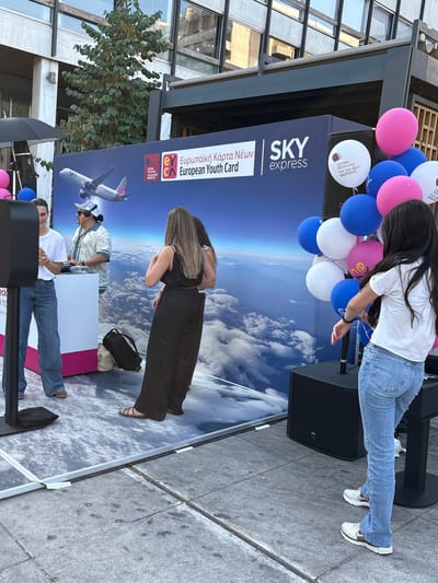 In this image, several people are gathered outdoors, seemingly at an event. The background features a large banner with the "SKY express" logo and the text "European Youth Card" in both Greek and English, suggesting a promotional or informational event related to youth travel or opportunities. Balloons, predominantly pink and blue, are present, adding a festive atmosphere.
One woman, wearing a white t-shirt and blue jeans, is bending over, possibly interacting with something near a speaker or stage. Another woman in a black dress is also seen from behind, with long brown hair. In the foreground, two men are visible. One, wearing a white t-shirt and blue jeans, is seated or leaning. Another man, wearing a light-colored patterned shirt and a baseball cap, stands behind a white counter.
The setting appears to be a public space, possibly a plaza or street, with buildings and trees visible in the periphery. The ground is paved with grey tiles, and the lighting suggests it is daytime, likely sunny. The overall mood seems lively and engaging, typical of a public event. The presence of promotional materials and the gathering of people indicate an ongoing activity. The text on the banner is clearly legible, providing context about the event's purpose.