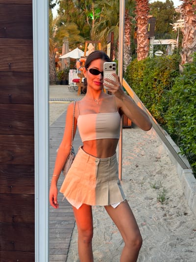A young woman is captured in a selfie taken at a beachside location in Bodrum, Turkey. She is wearing a light-colored, spaghetti-strap crop top and a light-colored skirt. She has on dark sunglasses and delicate jewelry, including a necklace and a bracelet. Her phone, with a textured white case, is held up to take the picture. The background shows sand, palm trees, and a wooden walkway. The sunlight suggests it is late afternoon or early evening, casting a warm glow on the scene. The overall atmosphere is relaxed and summery. The image is reflected in a glass surface, showing a glimpse of wooden panels below.