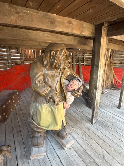 The image shows a man peeking out from behind a large wooden sculpture of a troll, located in the city of Satka, Russia. The troll sculpture is made of wood and depicts a large figure with a long beard and wearing a hat and skirt. The man is wearing a t-shirt and a baseball cap, and he is sticking his tongue out at the camera. The scene appears to be outdoors, possibly in a park or tourist attraction, with wooden planks forming the floor and walls. Red wooden panels are visible on the left side of the frame, and fishing nets are hanging in the background.