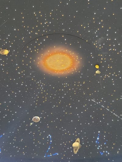 The image depicts a simulated night sky or planetarium dome. The central feature is a large, stylized sun with an orange and yellow glow. Scattered throughout the dark background are numerous stars of varying sizes and colors, primarily yellow and white. Several planets are visible, including a Saturn-like planet with prominent rings in the upper right, an Earth-like planet with a moon in the lower right, and what appear to be Mercury and Venus in the upper left. A thin, curved line is partially visible, likely representing the orbital path of a planet. Above and to the right of the sun is a string of closely spaced white dots, possibly representing a comet trail or a constellation. The image also depicts a blue constellation in the upper right corner of the shot. There is no evidence to support the image being from Reutov, Russia.