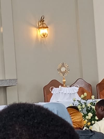 A brightly lit lantern hangs on a beige wall above a monstrance centered on a table adorned with white flowers. The monstrance sits between two wooden chairs with ringed backs, covered with a white cloth. The foreground is filled with the blurred heads of people attending what appears to be a church service.
