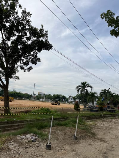This media file shows an outdoor scene at a construction site in Bundar, Indonesia. The image is oriented vertically, with the sky occupying the upper portion and the ground the lower. Several overhead power lines stretch across the frame.
In the foreground, a silhouetted tree with dense foliage is on the left. The ground appears to be a mix of dirt, gravel, and some patches of grass. Yellow and black striped caution tape runs along the edge of a pathway or excavated area. Several utility poles with lights are visible, one of which is in the mid-ground to the right, with a black base and a silver pole. A pile of trash and debris is scattered near this pole.
Further into the background, a large area of cleared and leveled earth is visible, suggesting ongoing construction. There are palm trees and other vegetation interspersed with buildings that appear to be houses or smaller structures, some with white exteriors. Construction equipment, specifically yellow bulldozers or similar machinery, can be seen in the mid-ground and further back. A tall light pole is also present in the distance. The sky is overcast with a mix of white clouds and a pale blue hue, indicating it is likely daytime, possibly morning or afternoon, with cloudy weather. There is no visible text in the image. The overall impression is one of a developing area, possibly a housing or commercial project, with elements of infrastructure and construction activity.