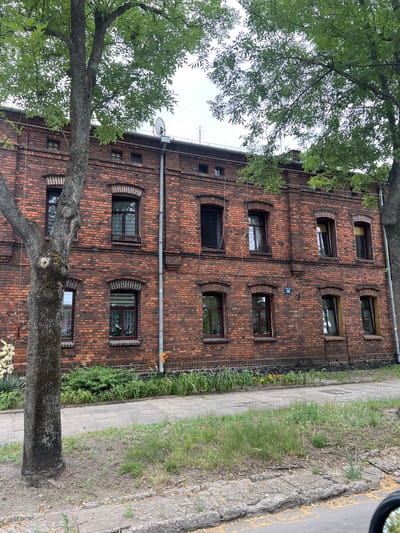 A brick building in Żyrardów, Poland, has a gray sidewalk in front of it. The building is two stories tall and made of red bricks with arched windows. Trees with green leaves are on either side of the building, and there are some plants in front of it. The sky is cloudy.
