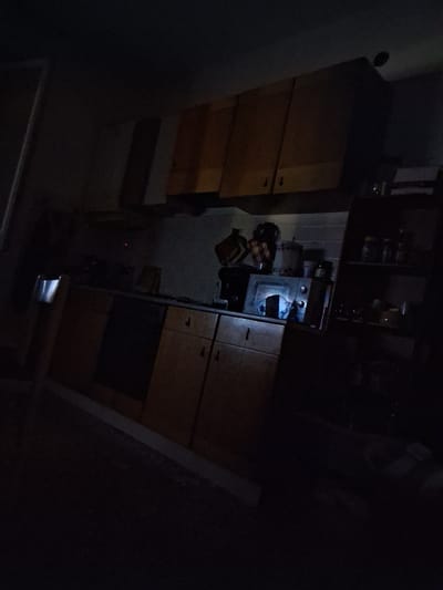 The image depicts a dimly lit kitchen, likely in an apartment in Colorno, Italy. The scene is largely obscured by shadow, but visible elements include wooden kitchen cabinets, a countertop, and various kitchen appliances and objects. In the center of the frame, a microwave oven is visible on the countertop, with a light emanating from within. To the right, a shelf unit holds several jars and containers. The overall impression is of a functional but somewhat cluttered kitchen, appearing undisturbed. No individuals are present in the image.