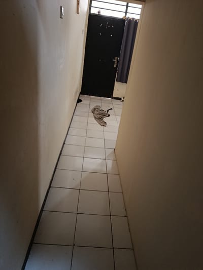 The image shows a narrow, tiled hallway. The walls are a neutral, off-white color, and the floor is covered in square, white tiles with dark grout lines. On the left wall, there is a light switch. At the end of the hallway, there is a black door with a silver handle and a small window at the top. To the right of the door, a dark grey curtain hangs. On the floor in the hallway, in front of the door, is a crumpled piece of light-colored fabric. The scene appears to be indoors, possibly in a residential building or a small commercial space, located in Depok, Indonesia. The lighting is dim, suggesting it might be late in the day or the area is not well-lit. There are no people visible in the image.