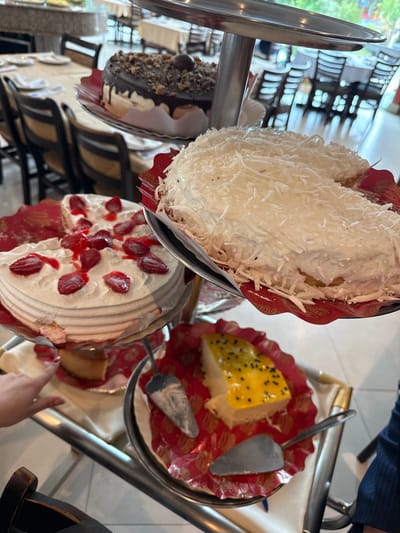 The image displays a vibrant selection of desserts arranged on a tiered stand, likely in a restaurant, buffet, or bakery setting in Rio de Janeiro, Brazil. The scene is well-lit by natural daylight, indicating it's daytime.
At the top of the stand is a large, round cake completely covered in white shredded coconut, resting in a decorative red paper liner. A serving utensil is positioned nearby. Below it, on the left, is a dark chocolate cake, generously topped with crushed nuts or crumbles and a chocolate drizzle.
On the right side of the middle tier, a white cake is adorned with fresh strawberry slices and a glistening red glaze or sauce; a portion has already been served, revealing the cake's interior. A metal serving spatula lies on its red patterned plate.
Further down, a slice of a bright yellow dessert, possibly passion fruit or mango, is visible, featuring distinctive dark seeds on its surface. This dessert also rests on a red plate. A person's hand, with neatly manicured nails, is seen reaching towards one of the desserts on the lower tier, suggesting they are about to serve themselves.
The background is softly blurred, showing parts of an indoor space with chairs and tables, and hints of outdoor greenery or reflections through a large window. Visible text on the red paper liners of the cakes reads "MR. BOLO," likely the brand name of the desserts. The overall impression is one of an abundant and appealing dessert spread.