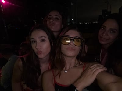 Four women are posing for a selfie in a dimly lit indoor location with a view of the Hong Kong skyline at night. The woman in the foreground is wearing large yellow-tinted glasses and a necklace with a heart pendant. To her left, another woman is making a kissing face. Behind them, a third woman is also making a kissing face. To the right of the woman in glasses, the fourth woman smiles toward the camera. Skyscrapers are visible through the window.