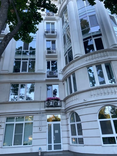 The media file is a vertical, eye-level photograph of a modern building facade with a mix of architectural styles. The building is predominantly light-colored, likely concrete or stucco, and features an array of windows. Some windows are large and rectangular, while others are smaller or even circular, forming part of a rounded architectural element on the upper floors. The building's facade is decorated with horizontal banding and a decorative curved molding with vertical embellishments.

Several air conditioning units are visible outside some of the windows, and one has a hanging planter with pink flowers. Trees with lush green foliage are visible in the foreground, with their branches extending into the frame from the left, partially obscuring some of the building's lower sections. Reflections of trees and sky are visible in the glass of the windows, suggesting it is daytime. The lighting indicates it is likely late afternoon or early evening, as the light appears soft and the sky is a muted blue. There are no people visible in the frame. The overall impression is of an urban architectural setting, possibly residential or commercial, with a touch of greenery. The location context is Nyzhni Mlyny, Ukraine, though this is not explicitly indicated by visual cues in the image itself. No discernible text is visible in the image.