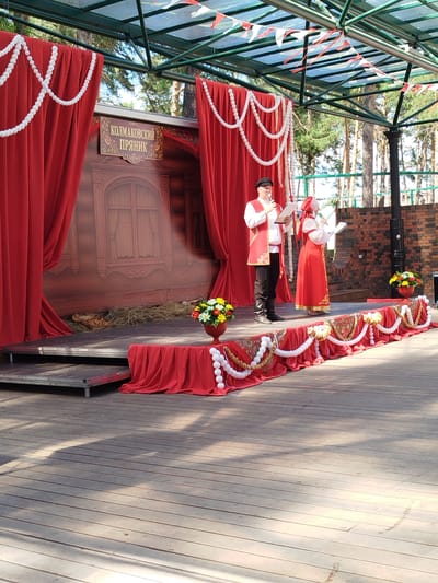 The image depicts an outdoor stage with a traditional Russian theme. Two individuals, a man and a woman, are standing on the stage, both dressed in traditional attire. The man wears a red vest over a white shirt, black pants, and a black cap. He holds a book or sheet of music and appears to be speaking into a microphone. The woman is dressed in a long red dress with intricate embroidery and a red headscarf. She is also holding a book and speaking into a microphone.
The stage is decorated with red curtains, white beaded garlands, and patterned fabric. Behind the performers, a backdrop depicts a rustic wooden house with a window. Hay is scattered on the stage floor in front of the backdrop. In front of the stage, a red cloth drapes over a decorated barrier, adorned with more white garlands and patterned fabric. Two vases of colorful flowers, one on the stage and one on the ground in front of the stage, add to the festive atmosphere.
The stage is set on a wooden boardwalk, and in the background, tall pine trees are visible under a bright sky. The overall impression is of a cultural performance or festival. Based on the provided information, this scene is located in the city of Zavodoukovsk, Russia.