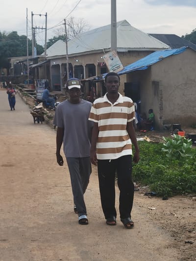 Two young men walk side-by-side down a dirt road in Madalla, Nigeria. The man on the left wears a grey t-shirt, grey pants, and black slide sandals with white stripes. He has a green and white baseball cap on. The man on the right wears a brown and white horizontally striped polo shirt and black pants, with black slide sandals. Behind them, to the left, is a woman in a blue dress walking away from the camera. Further back, a man is seated on a motorcycle. Buildings with corrugated metal roofs line the right side of the road, with one building displaying a large political poster of a man in a suit. Green foliage grows along the right edge of the road. The sky is overcast.