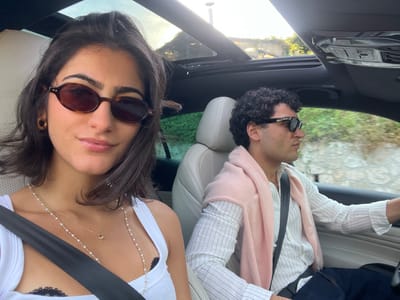 The image captures a candid, high-angle view of two individuals inside a car, likely enjoying a scenic drive in Èze, France, on a bright, sunny day. The perspective suggests the photo was taken from above them, looking downwards.

In the foreground, on what would be the bottom right when viewed upright, a woman with long, dark hair is visible, looking towards the camera. She wears dark, oval-shaped sunglasses and a white tank top, with a hint of black lace visible beneath. Delicate layered necklaces adorn her chest: one is a strand of small pearls, and the other a fine gold chain with a small heart pendant. A black seatbelt crosses her right shoulder.

Beside her, to the left, is a man seen in profile, looking towards the left. He has dark, curly hair and wears dark rectangular sunglasses. He is dressed in a light-colored, vertically striped long-sleeved shirt, with a soft pink garment, possibly a sweater or scarf, draped over his shoulders and chest.

The car's interior features light-colored leather seats and a dark roof lining. The top of the car appears to be open or has a large sunroof, revealing a clear blue sky and lush green foliage outside, indicating an outdoor, natural setting. The bright sunlight suggests pleasant weather. Both individuals appear relaxed and comfortable. No visible text is present in the image.