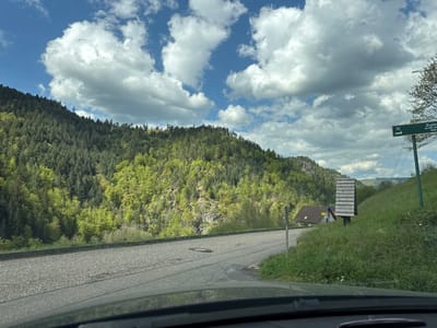 The image depicts a scenic mountainous landscape from the perspective of a vehicle, with a partial view of the dashboard at the bottom. The sky is clear blue with numerous fluffy white cumulus clouds. Dominating the middle ground are densely forested mountains, covered in a mix of light green deciduous trees and darker evergreen conifers, suggesting spring or early summer foliage. The terrain is rugged, with visible rock faces peeking through the trees on some slopes.
An asphalt road curves gently through the lower part of the frame, showing a manhole cover and a drain grate. To the right of the road, a grassy slope rises, featuring a tall, vertical wooden structure composed of horizontal slats, possibly an information board. Further up the slope and in the distance, a house with a dark brown roof is partially visible.
A green directional signpost stands on the right, displaying white text including "x1" and "Zum Große Sa..." with "400 m" beneath it, indicating a destination and distance. Faint power lines are also visible near the top right. The scene is bathed in bright daylight, suggesting good weather. The setting is a rural, natural environment, consistent with Forbach, Germany.