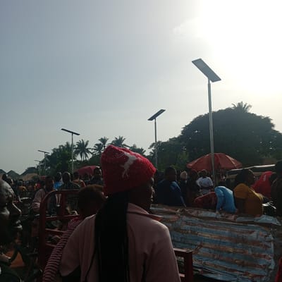 A densely populated outdoor scene in Nsukka, Nigeria, depicts a gathering of numerous individuals amidst market stalls and streetlights. The subjects are primarily African, with a variety of clothing styles and ages represented. Many individuals appear to be engaged in shopping or conversation. In the foreground, a woman wearing a red knit cap stands out. Several solar-powered streetlights are visible throughout the scene. Other trees are visible in the middle of the frame.