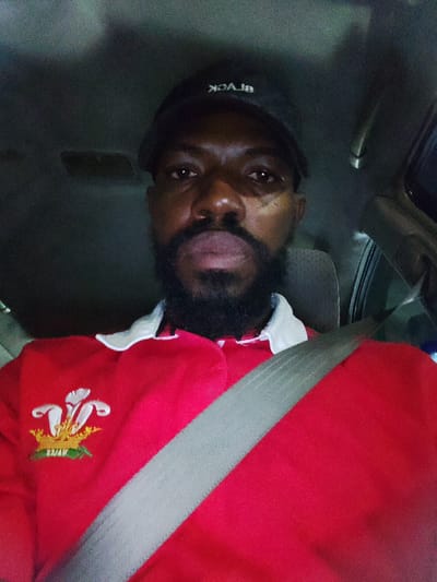 This is a selfie of a Black man sitting in the driver's seat of a vehicle. He is wearing a black baseball cap with white text that reads "BLACK" backwards. He also has on a red polo shirt with a white collar and a crest embroidered on the left side of the chest. The crest features a crown with a white feather plume and green foliage. The man has a beard and is looking directly at the camera with a neutral expression. A grey seatbelt is visible diagonally across his chest. The setting appears to be inside a car, possibly at night given the lighting conditions. The image quality is somewhat grainy, suggesting it may have been taken in low light. There are no immediate indications of the weather or a specific location beyond being in a vehicle. The image is cropped to focus on the man's upper body.