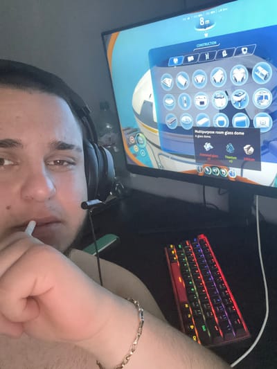 The image shows a person playing a video game on a computer. The game appears to be a construction or simulation type, as indicated by the "CONSTRUCTION" menu visible on the monitor. The player is seemingly building a "Multipurpose room glass dome" in the game. The computer setup includes a multicolored keyboard. The person has headphones on and is holding a cigarette in their hand. They are wearing a gold chain bracelet. There is no indication of Saedinenie, Bulgaria in the image.