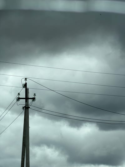 The image shows a concrete utility pole with multiple wires extending horizontally across a cloudy sky. The pole is positioned on the left side of the frame, with a crossbar supporting insulators and electrical wires. The sky is overcast with dark, heavy clouds, creating a somber atmosphere. A small, dark bird is visible in the upper right quadrant of the frame, flying against the clouds. The scene appears to be in an outdoor environment, possibly a rural or semi-urban area, given the utility pole and open sky. The location is specified as Mirny, Russia. There are no people or actions depicted in the image.