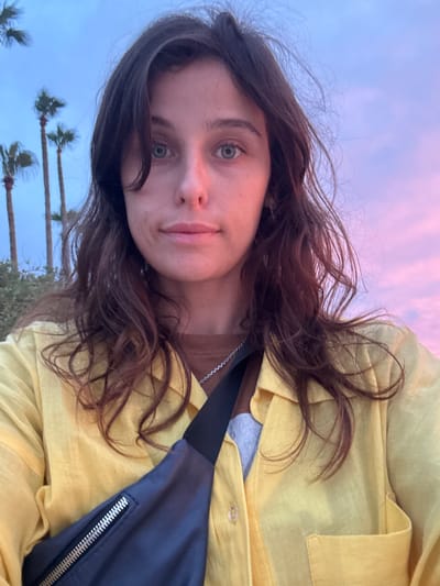 This is a close-up, slightly angled selfie of a young woman. She has long, dark brown, wavy hair that is partially obscuring her face. Her eyes are open and appear to be a greenish-blue color, and she is looking directly at the camera. Her lips are slightly parted, and she has a neutral expression. She is wearing a bright yellow collared shirt. Around her neck, a thin silver chain is visible. A dark blue fanny pack with a silver zipper is slung across her body, resting on the shirt.

The background suggests an outdoor setting, likely in the evening due to the soft, pastel colors of the sky, which transitions from pale pink to light blue. The presence of palm trees in the background indicates a warm climate. The image gives a sense of a relaxed, casual moment. There is no visible text in the image.