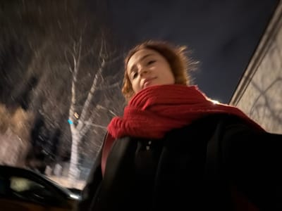 A woman in a black coat and large red scarf is standing outside at night. Bare trees and a building are visible in the background. The photo is taken from a low angle, making the woman appear taller. A car is partially visible in the lower-left corner. The image has some motion blur, indicating movement during the photo capture. The sky is dark.