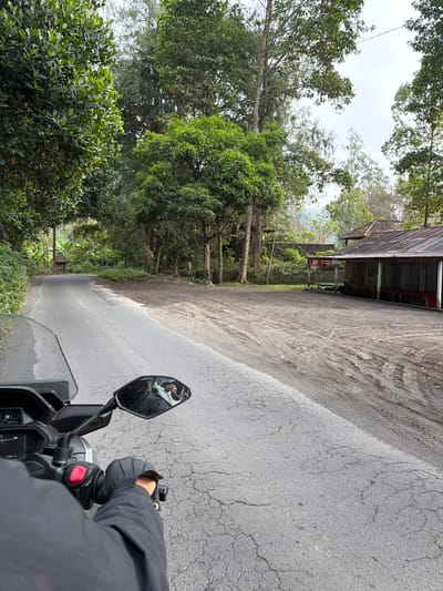 The image depicts a first-person view from a motorcycle traveling on a paved road in Kintamani, Indonesia. The road is cracked and shows signs of tire tracks from other vehicles. Lush green trees line the left side of the road, creating a canopy. In the distance, a small building with a red-tiled roof and open-air seating is visible on the right side of the road. A red and white sign is posted near the building. The sky is overcast, suggesting a cloudy day. The rider's hands are on the handlebars, and their reflection is visible in the motorcycle's side mirror, showing them wearing a helmet. The overall scene conveys a sense of travel through a tropical, rural environment.