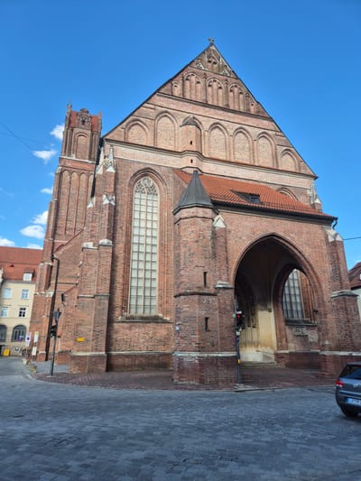 The image shows a large, red brick Gothic church, likely St. Martin's Church, in Landshut, Germany. The church has a tall, narrow tower on the left and a large, arched entrance on the right. The facade is adorned with arched windows and intricate brickwork. The street in front of the church is paved with cobblestones. A grey car is parked on the right side of the street, and a traffic light is visible near the entrance. The sky is blue with a few white clouds.