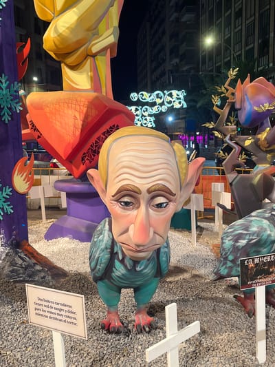 This image captures a nighttime outdoor scene featuring a large, cartoonish effigy that resembles Vladimir Putin with exaggerated features, including large ears and a prominent nose. The effigy is dressed in a teal, bird-like costume with red feet and talons. It stands on a gravel ground, surrounded by other colorful, oversized figures and decorations typical of a festival.
To the left of the central effigy, there's a purple structure adorned with a red flame motif and light blue snowflake shapes. Further back, a massive, yellow and orange structure looms, suggesting a larger, more complex art installation.
To the right, another effigy with dark, feathered wings and a long beak is partially visible. In front of this, a sign with dark text on a white background reads "LA MUERTE", and below that, "INVITADA A LA MESA DE ESTOS DOS".
In the foreground, a white sign with text in Spanish is placed on a stand. The text, in a decorative font, reads: "Dos buitres carroñeros tienen sed de sangre y dolor, pues los vemos muy enteros, mientras siembran el terror." This translates to: "Two scavenger vultures are thirsty for blood and pain, as we see them very whole, while they sow terror."
A white wooden cross is also visible in the foreground, placed on the gravel.
In the background, tall, modern buildings with lit windows suggest an urban environment. Streetlights and decorative light strings illuminate the scene, casting shadows and creating a festive atmosphere. The presence of construction barriers and orange fencing in the mid-ground indicates ongoing work or a designated event area.
This scene is highly characteristic of the "Fallas" festival, particularly the "Ninots" (large, artistic effigies) that are erected in the streets of cities like Valencia and Alicante, Spain. These effigies are often satirical and politically charged. Given the effigy's likeness to Vladimir Putin and the sign referencing "death" and "terror," it is a clear commentary on current geopolitical events. The overall impression is one of a public art installation designed for a festival.