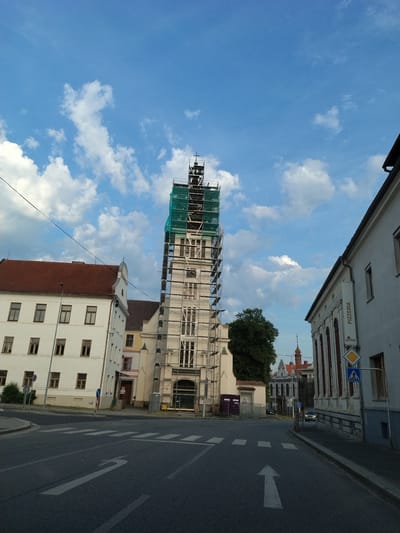 The image depicts a street scene in Horažďovice, Czechia, with a prominent bell tower undergoing renovation, covered in scaffolding and green netting. Several buildings line the street, including a white building with a red tiled roof to the left, and another white building to the right with a "PIZZERIA" sign. The street itself has crosswalks and directional arrows painted on the asphalt. The sky is blue with scattered white clouds. A few cars are visible on the road.