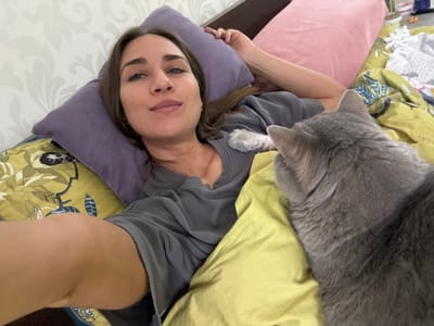 A woman is lying on her back in a bed with multiple pillows and blankets of various colors (yellow, purple, pink, and patterned). She is wearing a gray t-shirt. A gray cat is lying near her head. The location is Zanevka, Russia.