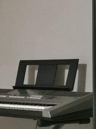 The image shows a close-up, eye-level view of a black keyboard stand positioned above a keyboard. The stand has a rectangular frame with two large openings. The keyboard is partially visible, showing white and black keys. The background is a plain, textured gray wall. The scene is indoors, likely in a residential setting, and there is no indication of the specific location within Bourg-lès-Valence, France, or any people present. The lighting appears to be from above and slightly to the left, casting shadows on the wall behind the stand.
