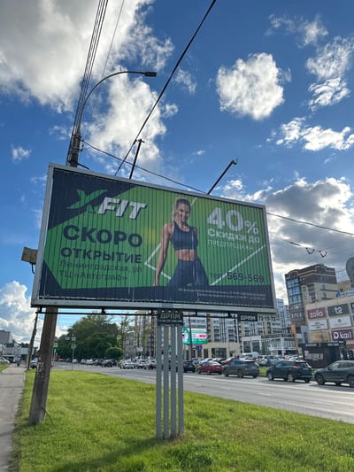 This is a medium-shot, eye-level, outdoor, daytime photograph of a large billboard advertising an X-Fit gym in Vologda, Russia. The billboard is green and white, with the X-Fit logo prominent at the top left. Below the logo, in large white letters, it reads "СКОРО ОТКРЫТИЕ" (Soon Opening). The address "Ленинградская, 98 ТЦ «Автограф»" (Leningradskaya, 98 TC "Autograph") is written below in smaller text. On the right side of the billboard, a woman in athletic wear is pictured, and the text "40% СКИДКИ ДО ПРЕДПРОДАЖА" (40% DISCOUNTS UP TO PRESALE) is displayed. A phone number, "тел. 569-569", is also visible.
The billboard is situated on the side of a road with multiple lanes of traffic. Several cars are driving on the road. In the background, there are modern buildings, including apartment complexes and commercial establishments with various logos like "Zolla", "LALE ANTILOP", and "Kari".
The sky is blue with fluffy white clouds, and there are overhead power lines and a street lamp visible. The foreground features a well-maintained grassy median strip and a sidewalk. The overall scene depicts an urban environment with commercial advertising.