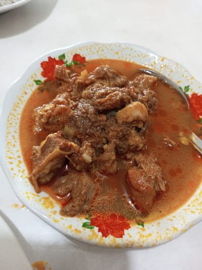 A plate of curry sits on a white surface. The curry appears to contain pieces of meat, potentially beef or lamb, and is covered in a reddish-brown sauce with oil droplets on the surface. The plate has a floral pattern of red roses around the rim. A spoon rests partially in the sauce. There is also a plate near the upper left corner of the picture, but it is out of focus.