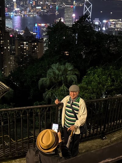 The image shows two people near a railing overlooking a nighttime cityscape, which is identified as Hong Kong, China. One person, wearing a cap, striped shirt, and light-colored jacket, stands near the railing and appears to be interacting with the other person. The second person, wearing a dark jacket and a tan hat, is holding a tablet. The cityscape includes illuminated buildings, a body of water with reflected lights, and structures such as the Bank of China Tower and the Hong Kong Observation Wheel. The location seems to be elevated, potentially a viewpoint or observation deck.