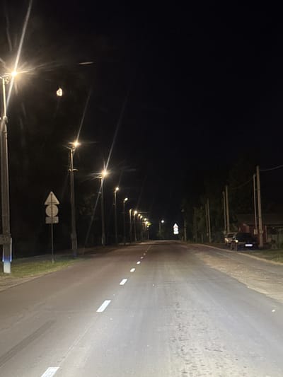 The image presents a nighttime scene in Лабжанскі сельскі Савет, Belarus, captured from a low, forward-facing perspective, likely from a vehicle on a paved road. The road is asphalt, featuring visible white dashed lane markings along its center. The surface appears somewhat damp or reflective in areas, and faint tire marks are present on the left side of the foreground.
A series of streetlights lines both sides of the road, extending into the distance. These lights emit a bright, warm-toned illumination, creating noticeable lens flare and starburst effects, particularly from the light sources on the left. Utility poles support these streetlights.
On the left side of the road, closer to the foreground, a post holds two stacked road signs: an upright triangular sign, white with a dark border, positioned above a circular sign, also white with a dark border. Beyond these, a grassy verge leads to dark, indistinct foliage.
The sky is dark and clear. In the upper-left quadrant, a small, luminous object with an irregular, textured appearance is visible. Above and slightly to the right of this object, a fainter, elongated streak of light is present.
On the right side of the road, a dirt or gravel shoulder is visible. Beyond this, dense, dark foliage and trees line the roadside. Further down the road on the right, several utility poles are present. A dark-colored sport utility vehicle or truck is parked off the road next to a structure, possibly a building or shed, which features a red-brown section. No individuals or animals are visible anywhere in the frame.