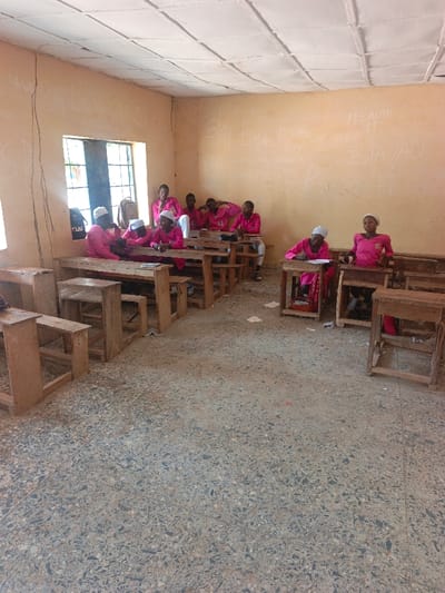 The image displays an interior view of a classroom or study area in Mada, Nigeria. Approximately ten individuals, predominantly young females, are present, uniformly dressed in pink long-sleeved tops; some also wear white or pink head coverings. They are seated at simple, unpainted wooden desks and benches arranged in rows.

The room features pale yellow walls extensively marked with white writing or graffiti. A single window with metal bars is visible on the left, admitting natural light and illuminating a black backpack and another bag resting near the window. The floor is a light-colored, worn terrazzo pattern with scattered paper debris. The ceiling consists of light-colored, possibly suspended, panels.

On the left side, a cluster of five individuals is seated, appearing to interact or observe their surroundings. Further right, individuals occupy separate desks; one person is actively writing on papers, and another in the front row is looking towards the viewer. Papers are visible on multiple desks and strewn across the floor.