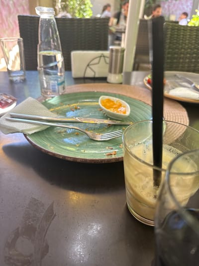 A messy table at an outdoor cafe. An empty glass with remnants of a smoothie and a black straw sits next to a green plate with remnants of food and a small dish containing orange sauce. A fork, knife, and crumpled napkin are on the plate. A bottle of water, a glass, and other obscured items are scattered on the table. In the blurred background, there are people and greenery suggesting an outdoor setting. Location unspecified but believed to be in Pernik, Bulgaria.