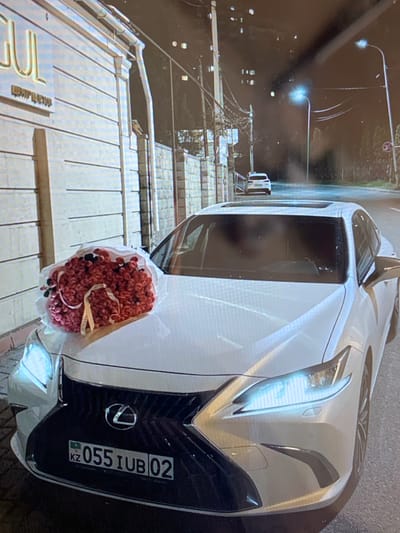 A white Lexus sedan is parked at night on a paved street, possibly in the city of Konstantinovka, Kyrgyzstan. A large bouquet of red flowers wrapped in clear plastic is placed on the hood of the car. The car has a license plate with the characters "KZ 055 IUB 02". In the background, a white SUV is visible further down the street. Streetlights illuminate the scene. A building with illuminated signage is to the left of the car. The overall impression is that of a celebratory or romantic gesture.